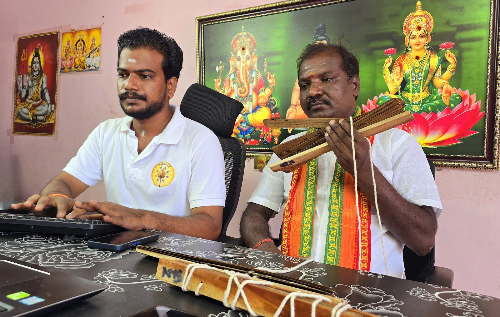 Nadi reader holding palm leaf bundles beside an assistant at a South Indian reading centre