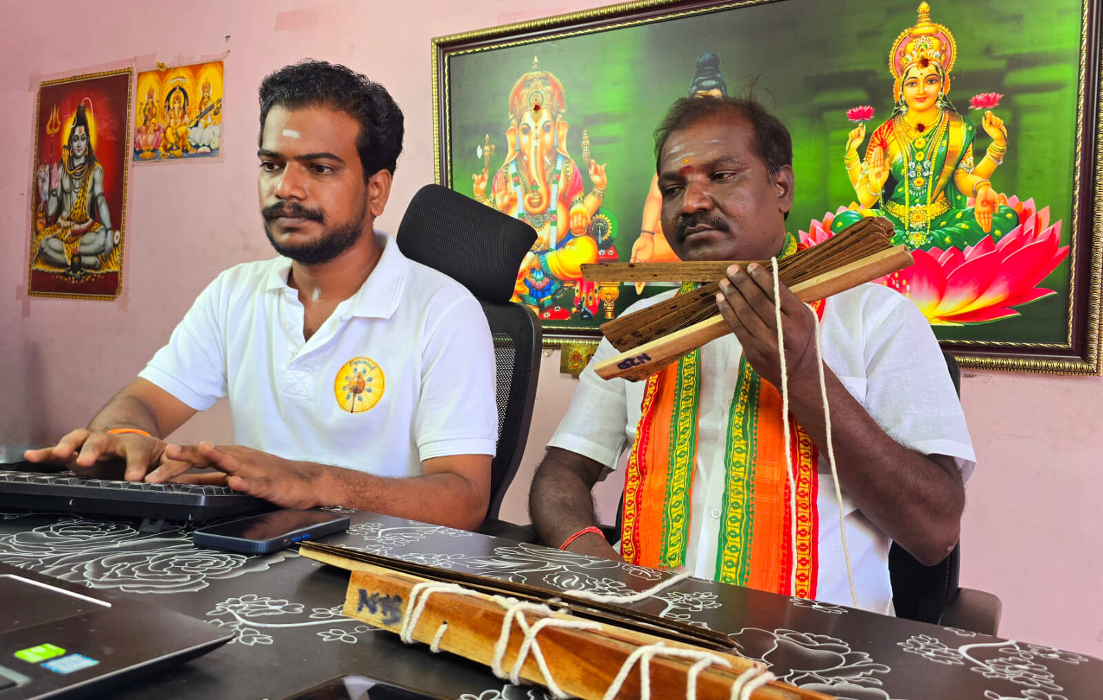 Nadi reader holding palm leaf bundles beside an assistant at a reading centre