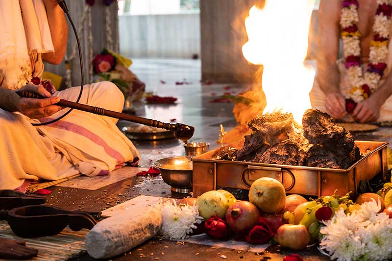 Hands offering ghee into the sacred homa fire surrounded by fruit and flowers