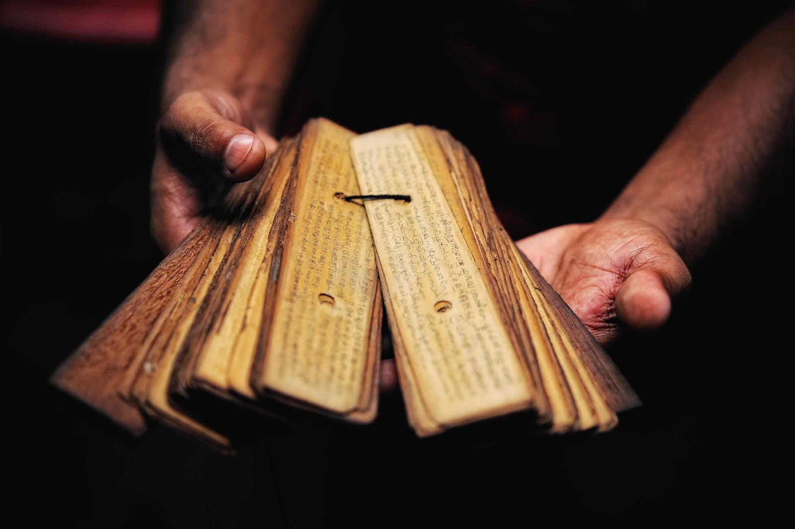 Hands holding an ancient palm leaf manuscript against a dark background